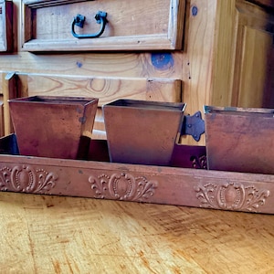 May include: A copper-colored planter with three square pots set in a rectangular tray. The tray has an ornate, embossed design along the front. The planter is made of metal and sits on a wooden surface.