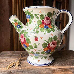 May include: Cream-colored ceramic pitcher with a spout and handle, adorned with hand-painted floral designs. The flowers are primarily pink and red roses, with green leaves and accents of blue and orange. The rim and base have blue trim.