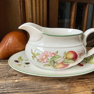 May include: White ceramic gravy boat and saucer set with a green trim and fruit and floral design. The gravy boat has a handle and spout. A wooden pear is to the left of the set.