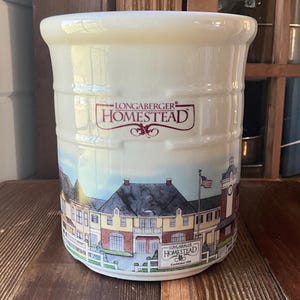 May include: Cream-colored ceramic Longaberger Homestead utensil holder. The cylindrical container features a detailed illustration of a building with a red roof, a clock tower, and a waving American flag. The Longaberger Homestead logo is displayed in red.