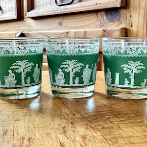 May include: Three vintage green and white glasses with a classical design. The glasses feature a green band with white figures, trees, and architectural elements. The rims have a decorative white pattern.