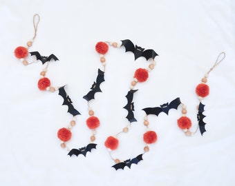 May include: A decorative garland featuring black paper bat cutouts, vibrant orange yarn pom-poms, and natural wooden beads strung on a jute twine cord. The garland includes loops at both ends for hanging and displays a repeating pattern of bats.