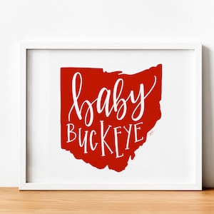 May include: A white framed print with a red outline of the state of Ohio. The text "baby buckeye" is written in white cursive lettering inside the outline.