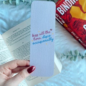 Puede incluir: Un marcap&aacute;ginas lila con esquinas redondeadas, con la inscripci&oacute;n "kiss all the time, disco occasionally" en escritura colorida. El marcap&aacute;ginas est&aacute; sobre un libro abierto y un libro rojo.