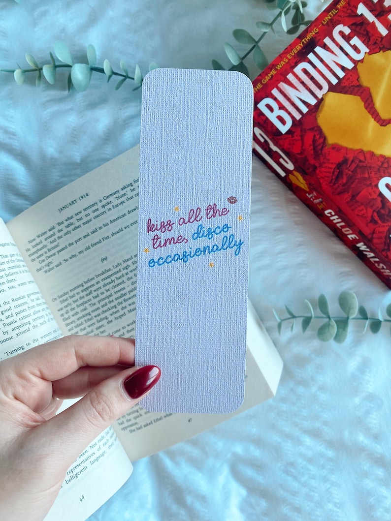 Puede incluir: Un marcap&aacute;ginas lila con esquinas redondeadas, con la inscripci&oacute;n "kiss all the time, disco occasionally" en escritura colorida. El marcap&aacute;ginas est&aacute; sobre un libro abierto y un libro rojo.