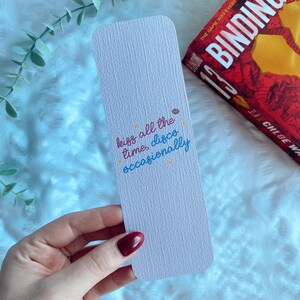 Puede incluir: Un marcap&aacute;ginas rosa claro con el texto "kiss all the time, disco occasionally" en escritura colorida. Cerca, se ve un libro con una portada roja y el t&iacute;tulo "Binding 13".