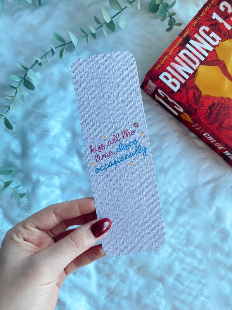 Puede incluir: Un marcap&aacute;ginas rosa claro con el texto "kiss all the time, disco occasionally" en escritura colorida. Cerca, se ve un libro con una portada roja y el t&iacute;tulo "Binding 13".