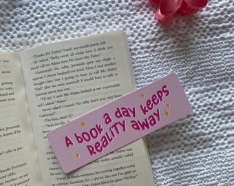 May include: A pink bookmark with the text "A book a day keeps Reality away" rests on an open book. A pink flower hair clip sits nearby. The book's pages are visible, with printed text.