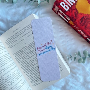 Puede incluir: Un marcap&aacute;ginas rectangular lila claro con el texto "kiss all the time, disco occasionally" en escritura colorida. Est&aacute; sobre un libro abierto, con un libro rojo en el fondo.