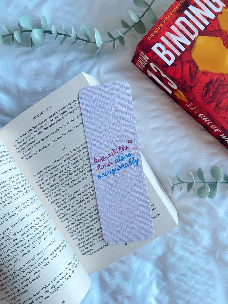 Puede incluir: Un marcap&aacute;ginas rectangular lila claro con el texto "kiss all the time, disco occasionally" en escritura colorida. Est&aacute; sobre un libro abierto, con un libro rojo en el fondo.