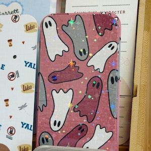 May include: A rectangular bookmark featuring a dusty rose background with a repeating pattern of white, grey, and dark red ghosts. The surface has a holographic star-shaped glitter finish that reflects light. The bookmark has rounded corners.