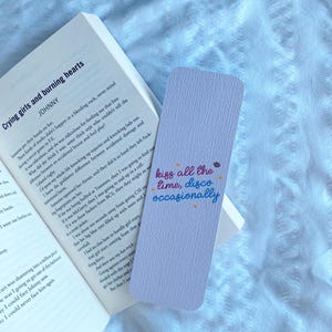 Puede incluir: Un marcap&aacute;ginas lavanda con el texto "kiss all the time, disco occasionally" en una fuente colorida y manuscrita. El marcap&aacute;ginas est&aacute; colocado en un libro abierto con el t&iacute;tulo "Crying girls and burning hearts".