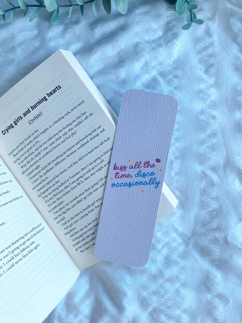 Puede incluir: Un marcap&aacute;ginas lavanda con el texto "kiss all the time, disco occasionally" en una fuente colorida y manuscrita. El marcap&aacute;ginas est&aacute; colocado en un libro abierto con el t&iacute;tulo "Crying girls and burning hearts".