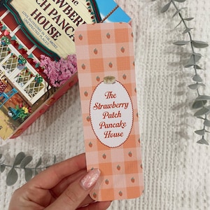 May include: A bookmark with a peach gingham pattern and small strawberry illustrations. The bookmark features the text "The Strawberry Patch Pancake House" in an oval design. A book with the same title is in the background.