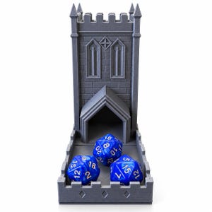 Puede incluir: Una torre de dados gris impresa en 3D con una estructura similar a un castillo, que presenta una torre alta y una entrada. Tres dados azules de veinte caras con números dorados se sitúan en la bandeja de la base.