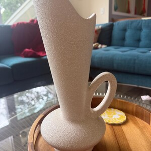 May include: A cream-colored ceramic vase with a textured, speckled finish. The vase features a bulbous base, a tall, flared neck with an asymmetrical rim, and a single curved handle attached to the side of the neck and the top of the base.