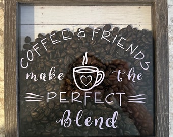 May include: A framed shadow box with a white background and a design of coffee beans, a coffee cup, and the text "Coffee & Friends make the perfect blend".
