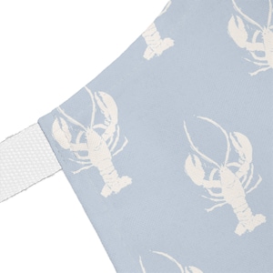 May include: A light blue apron featuring a repeating white lobster pattern. The fabric has a visible woven texture and includes a white fabric strap attached to the side corner, showcasing a clean, simple design with a consistent crustacean print.