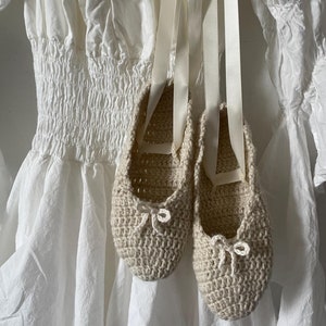 May include: A pair of beige crocheted ballet flats with white ribbon ties. The flats are hanging from a white dress with a ruffled hem.