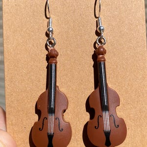 Peut inclure: Une paire de boucles d'oreilles en forme de violoncelle marron avec des crochets argentés. Les boucles d'oreilles présentent des détails noirs et blancs pour imiter les cordes et le corps d'un violoncelle. Fond marron.