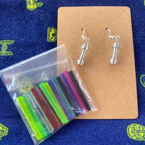 May include: A set of silver lightsaber earrings with a variety of colored lightsaber blades. The blades are made of plastic and come in a variety of colors, including green, blue, red, purple, and black.