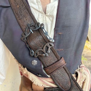May include: A dark brown leather baldric strap featuring an intricate embossed paisley pattern and a large, ornate, dark metal buckle. The strap is worn over a dark gray vest and a cream-colored shirt, with a tan leather belt visible below.
