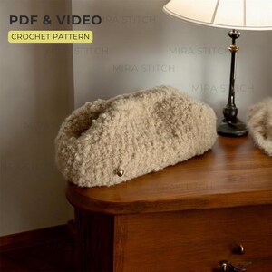 May include: A beige crochet clutch bag with a textured, fluffy appearance. The bag has a small gold-colored heart-shaped embellishment. The image also includes text that reads "PDF & VIDEO CROCHET PATTERN".