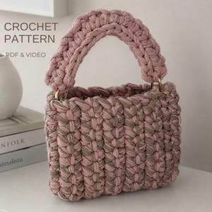 May include: A crochet pattern PDF and video for a handbag made of thick, dusty pink and taupe yarn with metallic gold thread accents. The bag features a rectangular body with a thick, rounded top handle attached by gold-coloured metal rings.