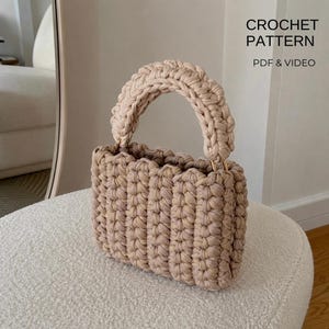Op de afbeelding: Een beige gehaakte handtas met een dik, gevlochten handvat en een gestructureerd ribbelpatroon. De tekst CROCHET PATTERN PDF & VIDEO is zichtbaar in de rechterbovenhoek. De tas heeft een rechthoekige vorm met afgeronde hoeken.