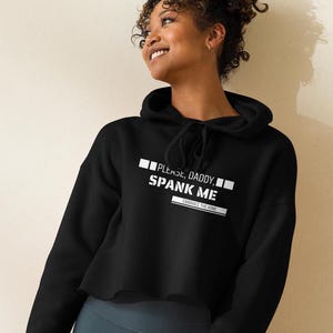 Puede incluir: Sudadera corta negra con el texto "PLEASE, DADDY, SPANK ME" en letras blancas. La frase "EMBRACE THE KINK" está debajo. La sudadera con capucha tiene un cordón y la lleva una persona.