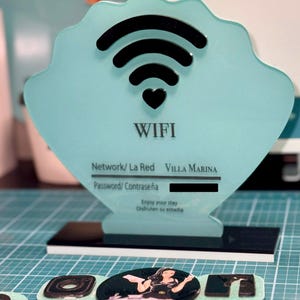 Op de afbeelding: Een turquoise blauwe schelpvormige bord met een zwart wifi-symbool en een hart. Het bord heeft de tekst "WIFI", "Network/ La Red", "VILLA MARINA", "Password/ Contraseña", "Enjoy your stay" en "Disfruten su estadia".