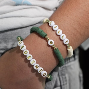 Peut inclure: Trois bracelets sont présentés sur un poignet. Un bracelet épelle "REDEEMED" en lettres dorées sur des perles blanches. Un autre bracelet épelle "LILLY" en lettres dorées sur des perles blanches. Un troisième bracelet est vert avec des accents dorés.