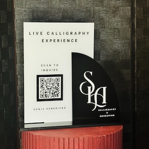 Può includere: Un cartello in bianco e nero con il testo "LIVE CALLIGRAPHY EXPERIENCE" e "SCAN TO INQUIRE". Il cartello presenta anche un codice QR e il nome "SONJA HENDRICKS". Un semicerchio nero mostra le lettere "S & H" e le parole "CALLIGRAPHY & ENGRAVING".