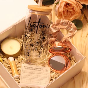 May include: A gift box with a glass tumbler, candle, lip balm, compact mirror, and scrunchie. The tumbler has a floral design and the name "Victoria". The box is filled with shredded paper and decorated with flowers.