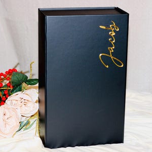 May include: A black gift box with a gold script name "Jacob" on the front. The box is rectangular and has a matte finish. The background includes flowers and greenery.