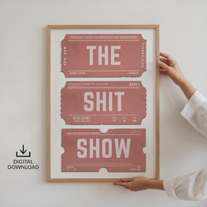 Può includere: Una stampa incorniciata con tre biglietti in stile vintage in una tonalità rosa polveroso, ognuno con una parola in testo bianco in grassetto: "THE", "SHIT" e "SHOW". La stampa è in una cornice di legno chiaro. Il testo "DIGITAL DOWNLOAD" è in basso a sinistra.