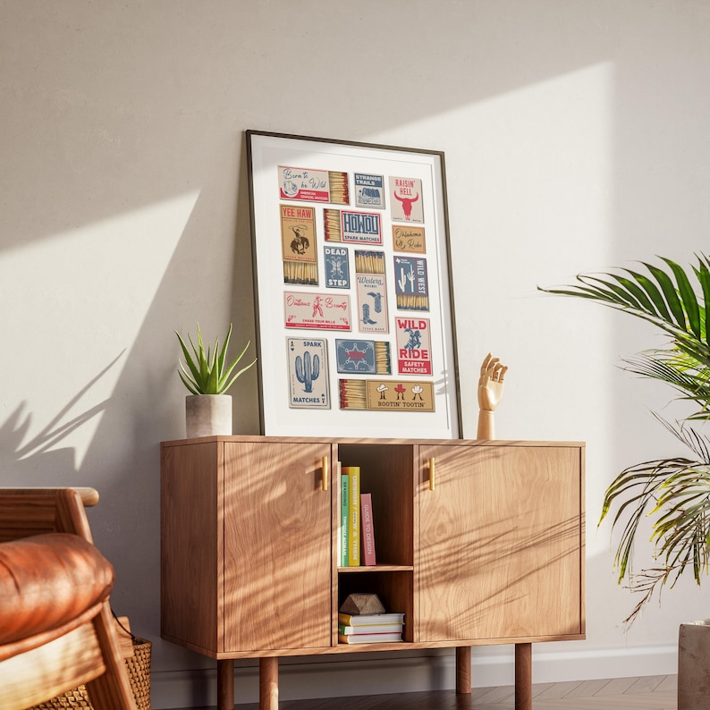 A eclectic print displaying retro matchbook designs with americana themes. The artwork includes illustrations and text, such as cacti, boots, and the words "Born To Be Wild" The print is displayed on a wooden cabinet.
