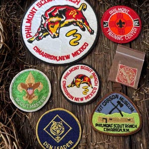 May include: A collection of embroidered patches featuring text and graphics. The patches include the words "Philmont Scout Ranch," "Cimarron, New Mexico," "Training Center," "Kit Carson Museum," and "Den Leader." The patches are in various colors.