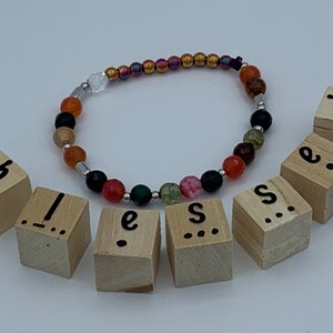 May include: A beaded bracelet featuring a mix of colourful stones and a silver charm. The bracelet is spelled out with wooden blocks that spell "blessed".