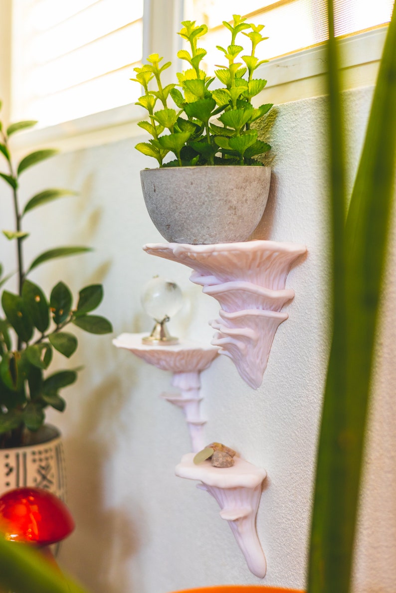 Op de afbeelding: Een set van drie witte, paddenstoelvormige wandplanken met een kleine plant in een pot op de bovenste plank, een glazen bol op de middelste plank en een kleine steen op de onderste plank.