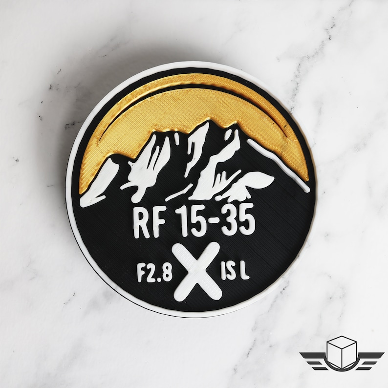 Pu&ograve; includere: Un cerotto rotondo nero e oro con un design a catena montuosa bianca. Il testo "RF 15-35 F2.8 X IS L" &egrave; stampato sul cerotto.