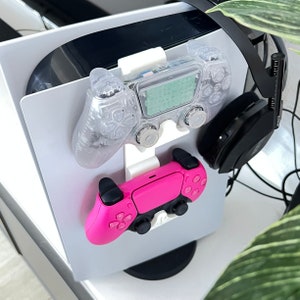 May include: A white 3D printed stand with two video game controllers and a pair of black headphones. One controller is clear and the other is pink.