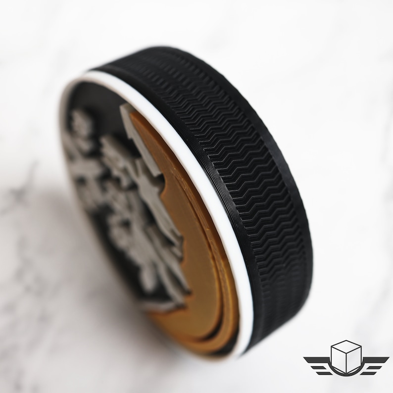 Pu&ograve; includere: Un disco da hockey nero e bianco con un design dorato e grigio. Il disco ha un bordo bianco e una superficie testurizzata.