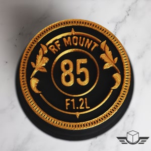 Pu&ograve; includere: Un tappo per obiettivo della fotocamera rotondo nero e oro con la scritta "RF MOUNT 85 F1.2L" stampata su di esso.