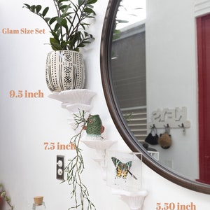 Op de afbeelding: Set van drie witte keramische wandplanken in de vorm van een paddenstoel. De planken zijn 9,5 inch, 7,5 inch en 5,5 inch lang. De planken zijn versierd met een plant in een pot, een klein uilbeeldje en een ingelijste vlinder.