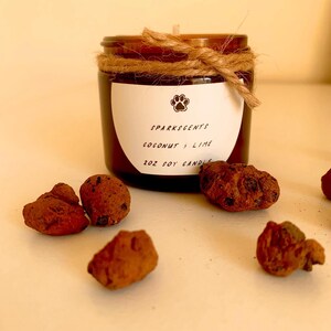 Peut inclure: Une bougie en verre brun dans un bocal avec une étiquette blanche sur laquelle est inscrit "SPARKSSCENTS COCONUT & LIME 2OZ SOY CANDLE". Le bocal est attaché avec de la ficelle et porte un logo en forme de patte sur l'étiquette.