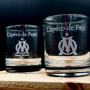 Peut inclure: Deux verres à whisky transparents avec des motifs gravés. L'un porte l'inscription "L'apéro de Papa" et l'autre "L'apéro de Papi". Les deux présentent un emblème argenté et le texte "DROIT AU BUT". Les verres sont posés sur une surface en bois.