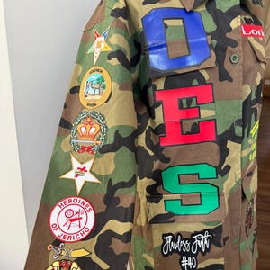 May include: Camouflage jacket with patches and lettering. The jacket features the letters "DES" in blue, red, and green, along with various patches on the sleeves. The text "Flawless Faith #40" is also visible.