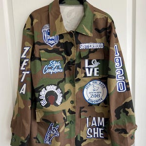 Peut inclure: Une veste utilitaire de couleur camouflage vert avec divers patchs, dont "ZETA", "Style & Confidence", "SISTERHOOD", "LOVE", "BLACK GIRL MAGIC", "ZETA PHI BETA", "ZΦΒ SORORITY", "I AM SHE", et "1920".