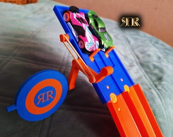 Könnte beinhalten: Ein blauer und orangefarbener Spielzeug-Autostarter mit einem Gummiband und zwei Spielzeugautos. Die Autos sind ein pinkfarbenes und weißes Auto und ein grünes Auto. Der Starter hat eine kreisförmige Zielscheibe mit den Buchstaben "AR" darauf.